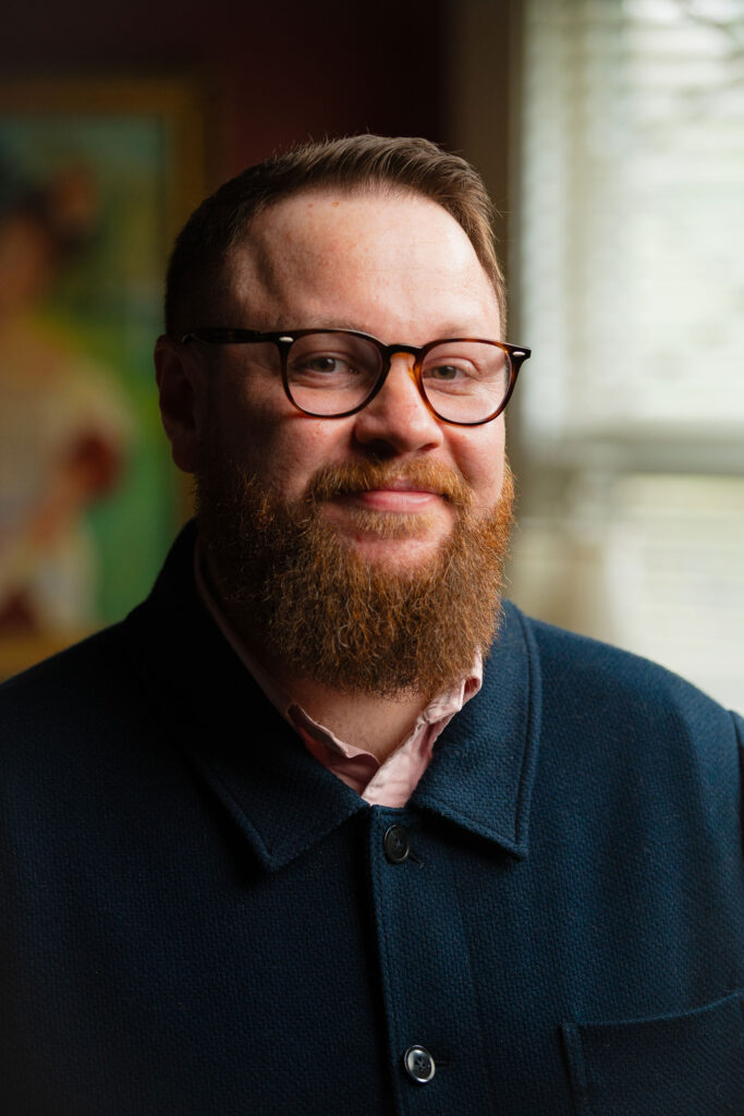 A man with short brown hair and a reddish-brown beard stands by a window indoors, smiling. He is wearing brown-rimmed glasses and a light pink collared shirt underneath a textured navy button-down shirt.
