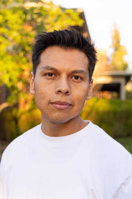 Chris Hoshnic, a Diné poet, stands outdoors, looking at the camera with a small smile on his face. He wears a white tee shirt. In the background is a sunlit tree and a house.