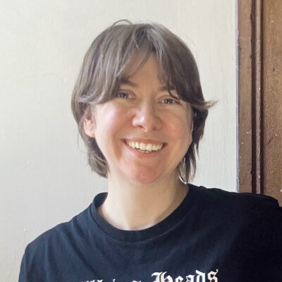 Kate Pyontek, a person with brown wavy hair that reaches the back of their neck, stands against a wall and wooden doorframe and smiles at the camera. They are wearing a dark tshirt with white lettering on it.