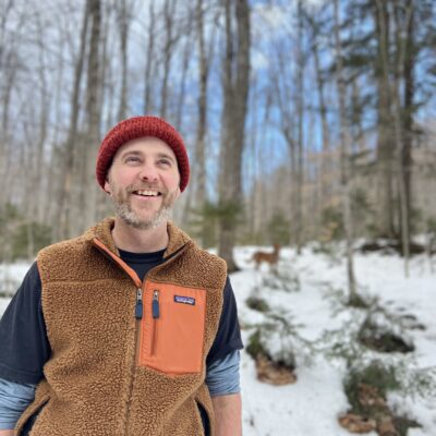 The poet Michael Garrigan stands in a snowy forest landscape, smiling and looking up at the sky. He is wearing a red beanie, a brown and orange hiking vest, and has a brown beard stippled with white.