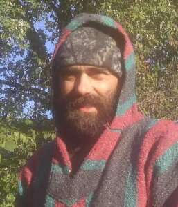 A man wearing a felted red, teal, and grey hooded shirt and a dark grey beanie stands outdoors near a large tree. He has a thick beard.