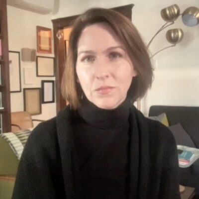 The poet Rebecca Morton sits indoors, looking at the camera. She wears a black turtleneck sweater and has brown hair. In the background is a bookshelf, two couches, a lamp, and frames on the wall.