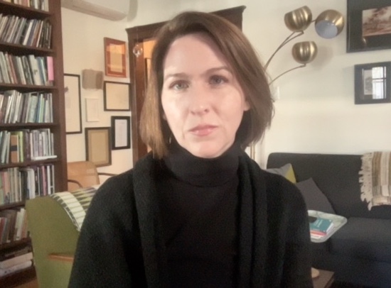 The poet Rebecca Morton sits indoors, looking at the camera. She wears a black turtleneck sweater and has brown hair. In the background is a bookshelf, two couches, a lamp, and frames on the wall.
