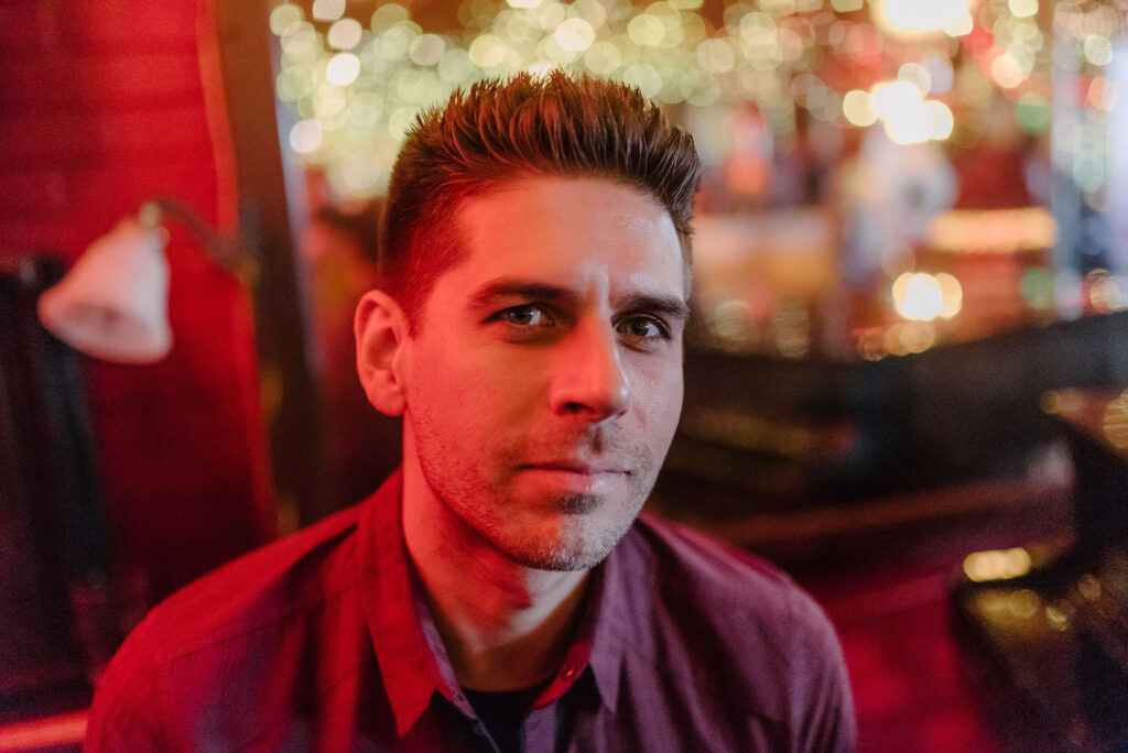 Carlos Andrés Gómez, a man with spiked hair, under a red-tinted glow, wearing a red, button-down short-sleeved shirt, looks at the camera on a three-quarter angle.