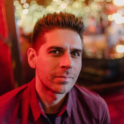Carlos Andrés Gómez, a man with spiked hair, under a red-tinted glow, wearing a red, button-down short-sleeved shirt, looks at the camera on a three-quarter angle.