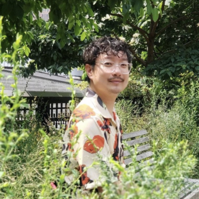 Chen Chen sits on a bench surrounded by greenery. He wears glasses with clear frames and a short-sleeved shirt with a bright orange floral pattern. Photo Credit: Anna Jekel.