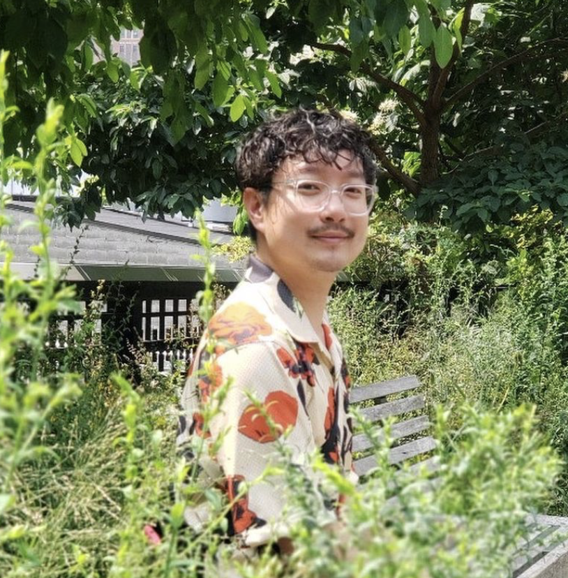 Chen Chen sits on a bench surrounded by greenery. He wears glasses with clear frames and a short-sleeved shirt with a bright orange floral pattern. Photo Credit: Anna Jekel.
