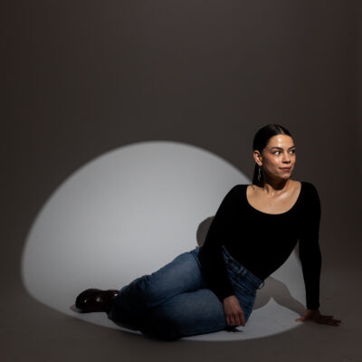 The poet Cheyenne C. Fletcher sits on the ground indoors under a spotlight, leaning on one arm and smiling off to the side. Her long black hair is pulled back into a ponytail and she wears a fitting long-sleeved black scoop-neck top, blue jeans, and dangling earrings.