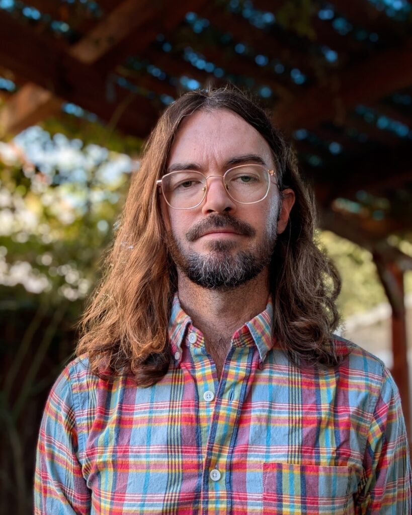 Nicholas Yingling stands outdoors under a wooden structure, looking at the camera with a neutral expression on his face. He wears wire-rimmed glasses and a colorful button-down checkered shirt. He has a beard, thick dark brown eyebrows, and wavy brown hair that falls to his shoulders.