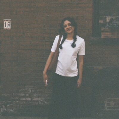 The poet Rukan Saif stands outdoors smiling against a brick wall on a street pavement. Her black hair is in braids and she wears a white tee shirt, a long black skirt, black Mary Jane shoes, and white socks with printed hearts. She is carrying a brown shoulder bag and carrying a phone in one hand.