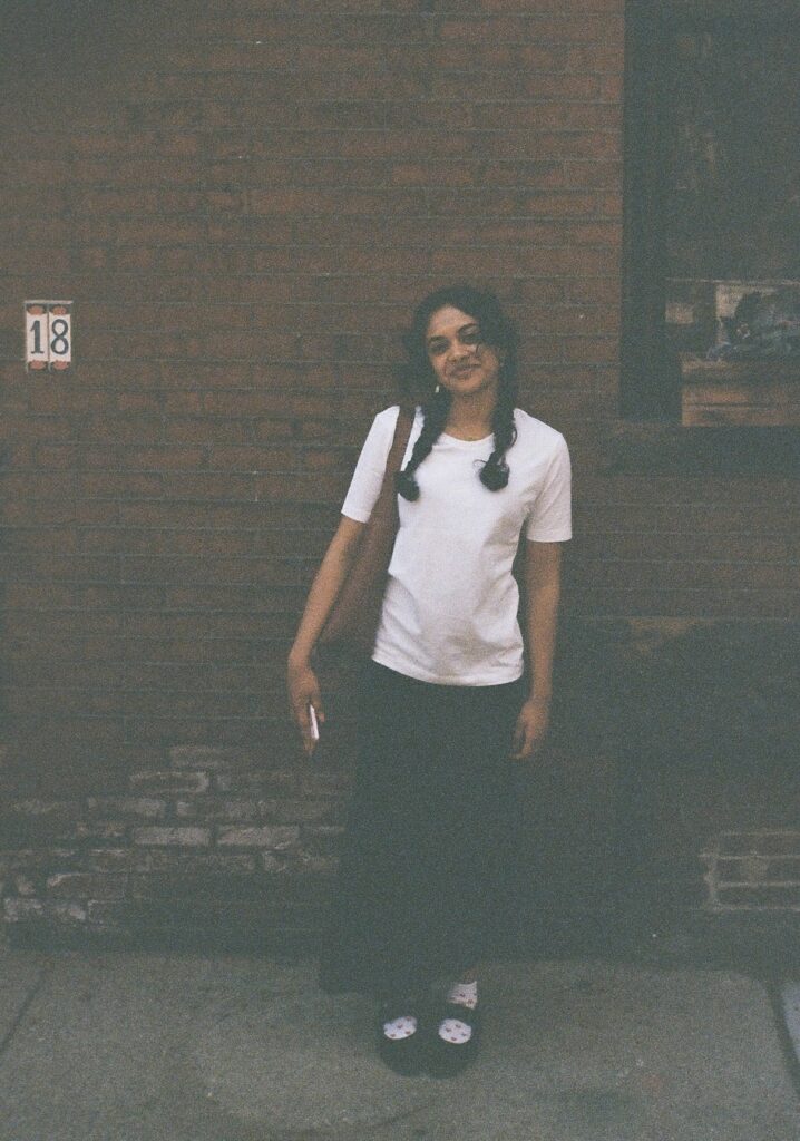 The poet Rukan Saif stands outdoors smiling against a brick wall on a street pavement. Her black hair is in braids and she wears a white tee shirt, a long black skirt, black Mary Jane shoes, and white socks with printed hearts. She is carrying a brown shoulder bag and carrying a phone in one hand.