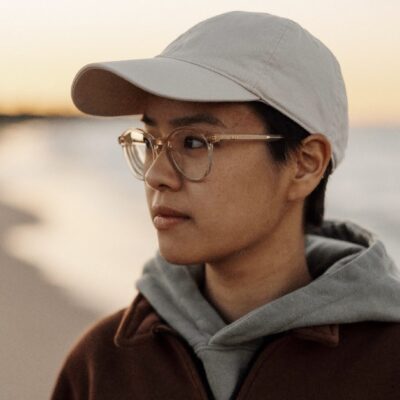 Eliana Chow, an Asian American poet wearing clear light brown round glasses, a beige baseball cap, brown collared fleece jacket, and grey hoodie, stands by the ocean, looking off to the side.