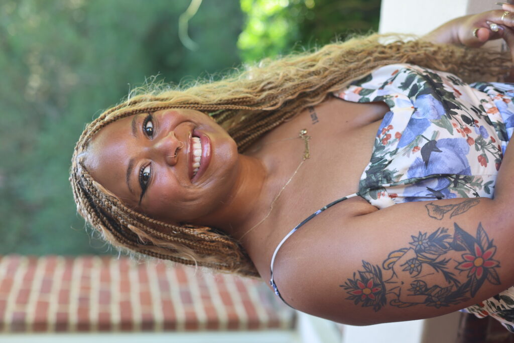 February Spikener, a Black femme poet, sits outside with a brick wall and a garden in the background. She has tattoos and is wearing a floral dress.