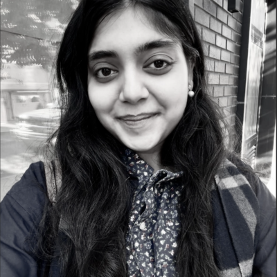 Vasvi Kejriwal, a young Indian woman, stands outside on a street, smiling at the camera. She wears a dark jacket, plaid scarf, and dark button-down shirt with flowers printed on it. There is a window in the background reflecting a passing car and a tree. Next to the window is a brick wall.