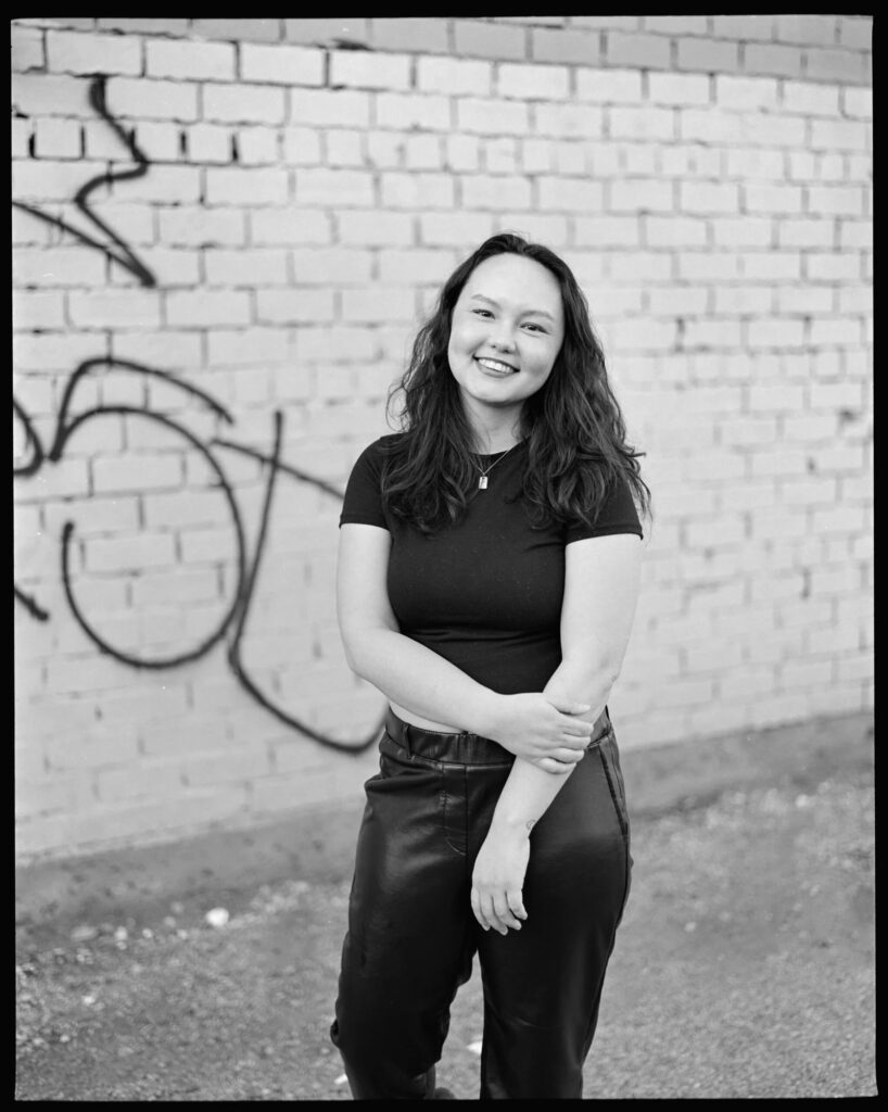 Arah Ko stands outdoors in front of a brick wall with graffiti on it. She is smiling and wears a small pendant on a silver chain, black pants, and a black tshirt.