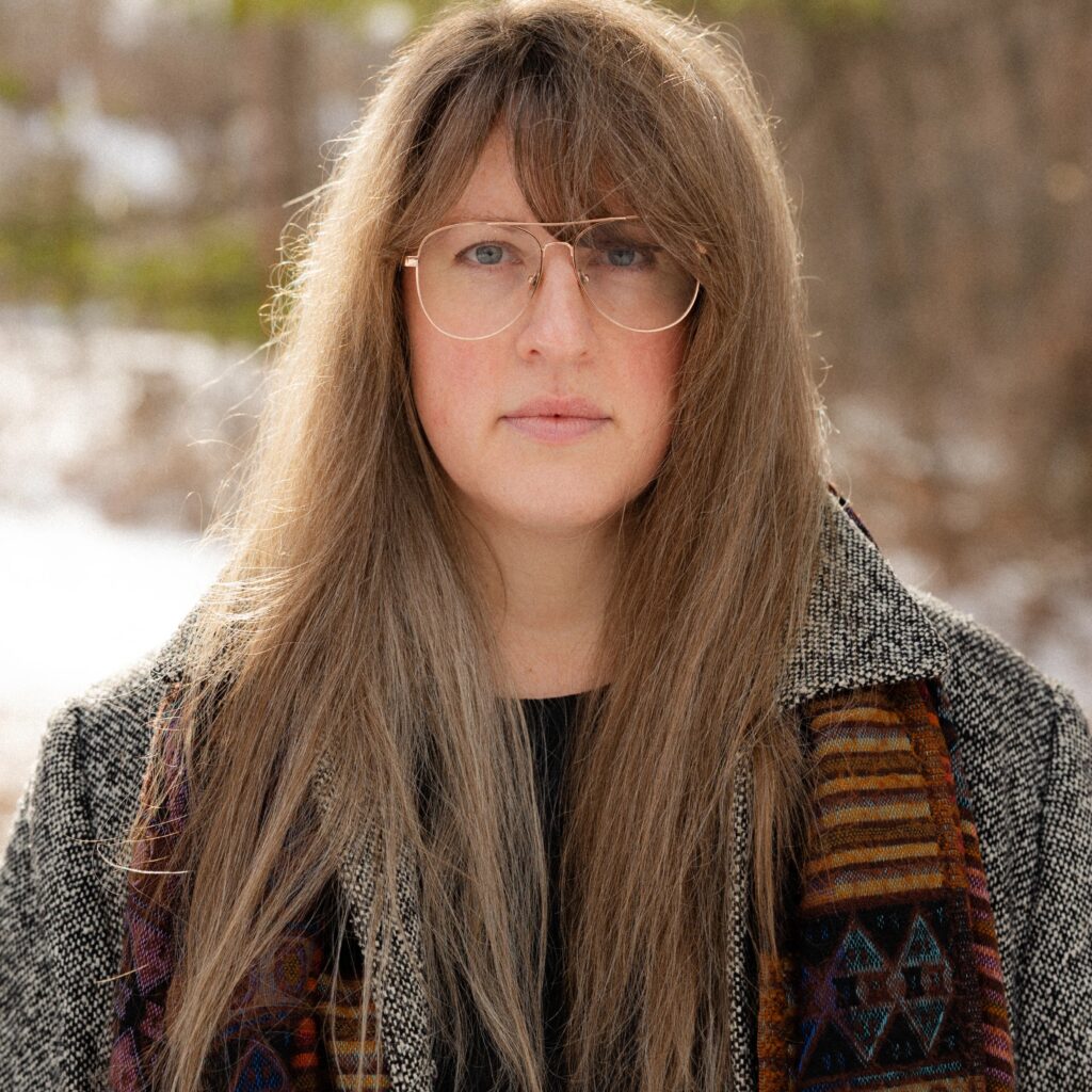 Meg Reynolds stands outdoors in a winter landscape, looking at the camera. She wears large wire-rimmed glasses and has long, light brown hair and grey eyes. She is wearing a woven scarf and a tweed coat.