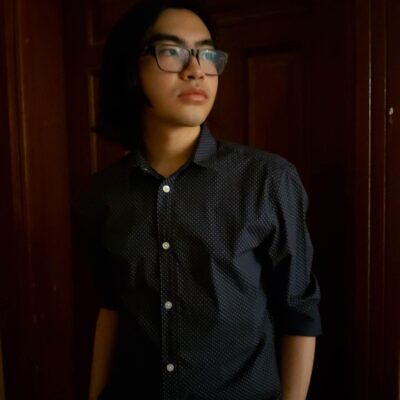 The poet MJ Gomez, a young Filippino poet, stands in a dark space, looking off to the side. He wears dark thick-framed glasses and a dark short-sleeved button-down shirt with pearl buttons. He has thick eyebrows and a faint moustache.