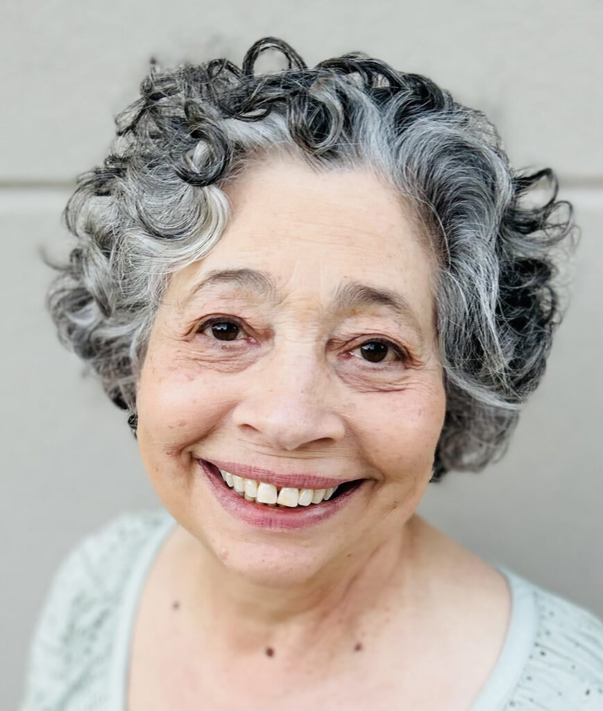 A woman with curly black, gray, and white hair and dark brown eyes smiles at the camera. She wears a pale green blouse.