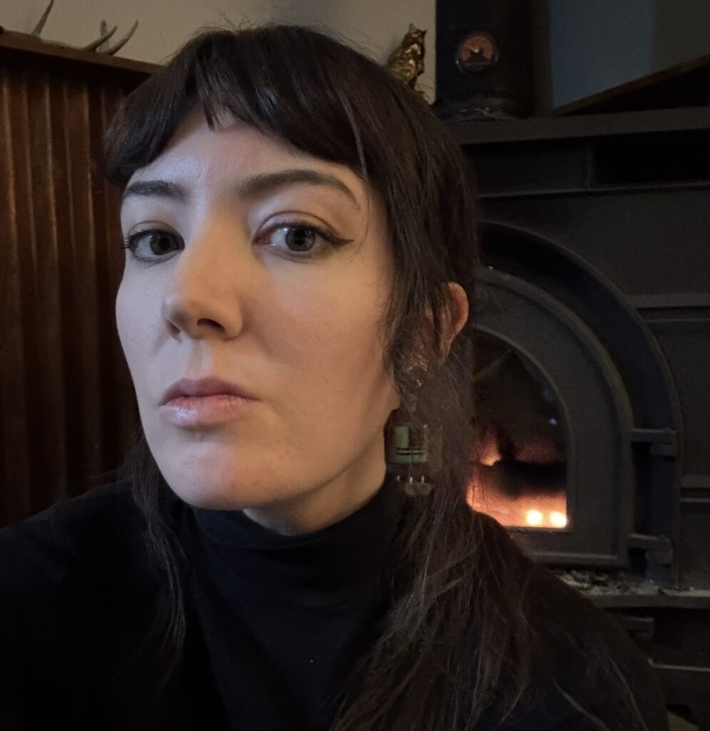 A young woman with dark brown hair and dark brown eyes looks at the camera. She is wearing a black turtleneck sweater and dangling earrings. A lit fireplace appears behind her.