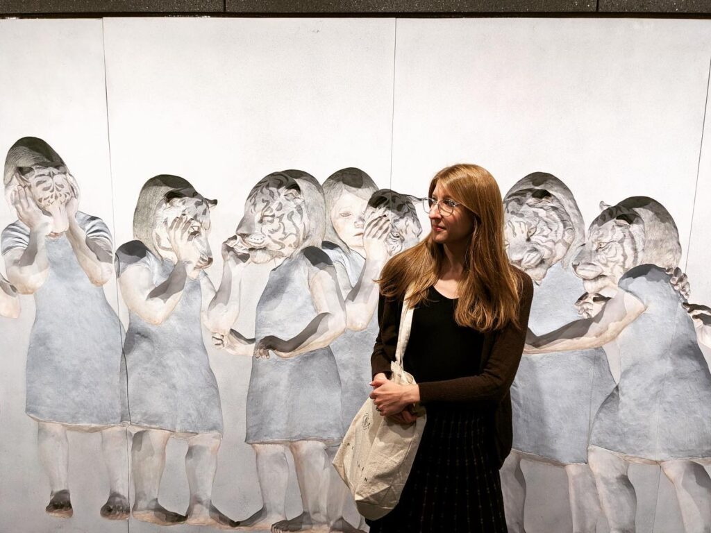 Catherine, a woman with long blonde hair and blue glasses, looks to the side at a pastel blue and gray mural of children wearing tiger masks.