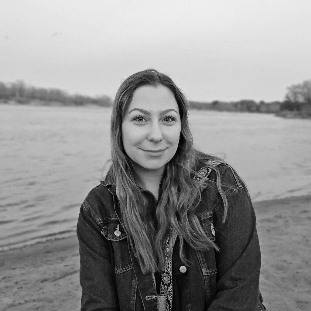 A young woman with long hair stands outdoors near a body of water. She wears a denim jacket and is smiling at the camera.