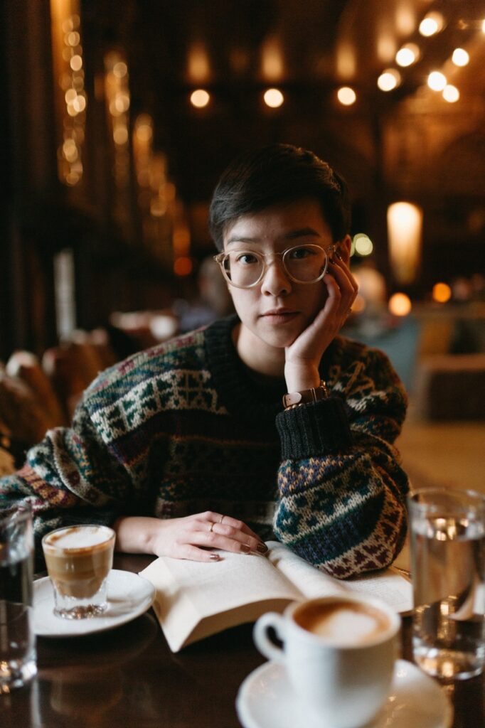 Eliana Chow, a young Asian woman with short black hair, looks up from a book in a cafe with dark walls and warm light. She is wearing a knit sweater with dark green and white patterns and wears round glasses and two rings on her fingers. Her fingernails are painted a dark reddish-brown and there is a cortado beside her.