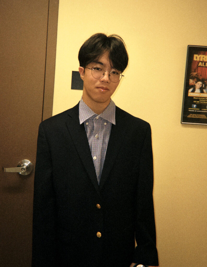 Giljoon Lee is standing indoors against a yellow wall, wearing round glasses, a dark blazer, and a checkered button-up shirt. To the left is a closed door, and a colorful poster is partially visible on the right. The lighting is warm.