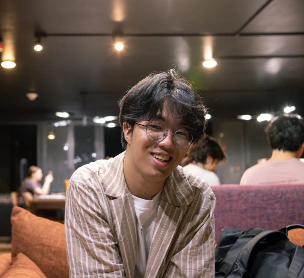 Poet Giljoon Lee, a young Korean man with short black hair, sits on a couch in a room with others studying in the background. He wears glasses and a long-sleeve button-down shirt open to a white tshirt. There is a backpack beside him and he is smiling.