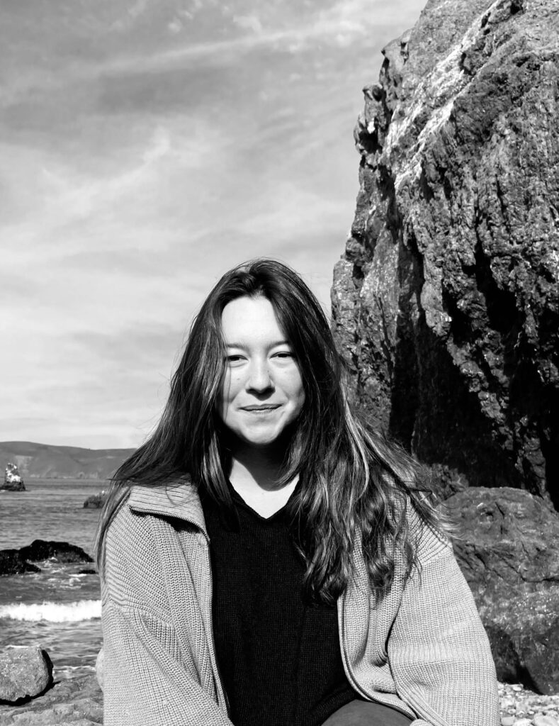 black and white photo of author (woman with long hair) at the beach. Wearing a sweater, slight closed mouth smile, cliff and water in the background