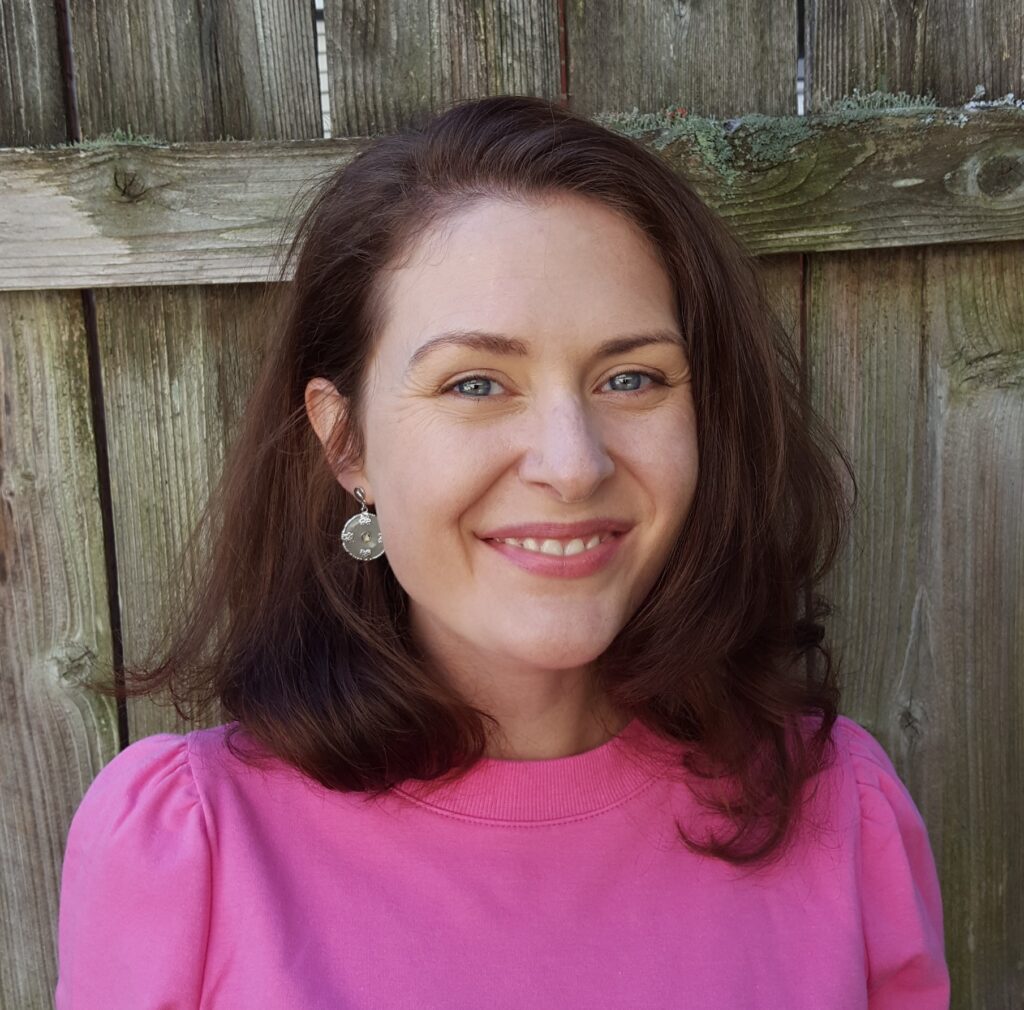 Mallory Rodenberg, a poet with wavy, shoulder-length brown hair and blue-gray eyes, smiles at the camera. She wears pink clothing cinched at the shoulders and dangling earrings. There is a wooden fence behind her.