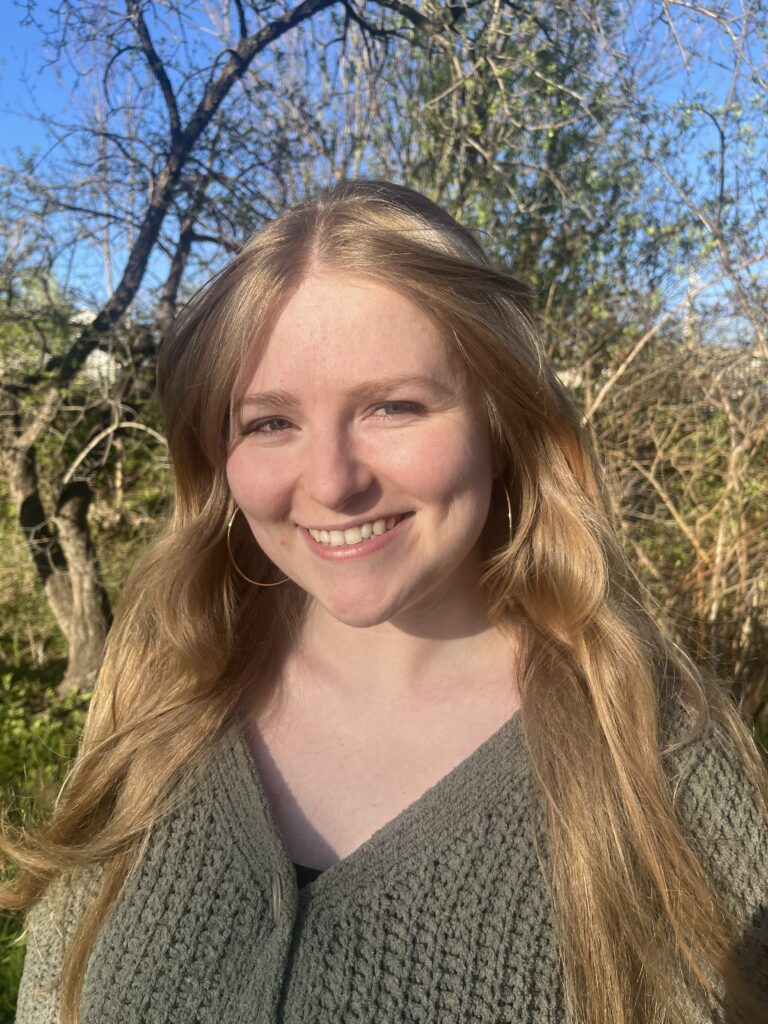 A smiling woman with long blonde hair standing outdoors, wearing large hoop earrings and a green sweater, with trees and blue sky in the background.