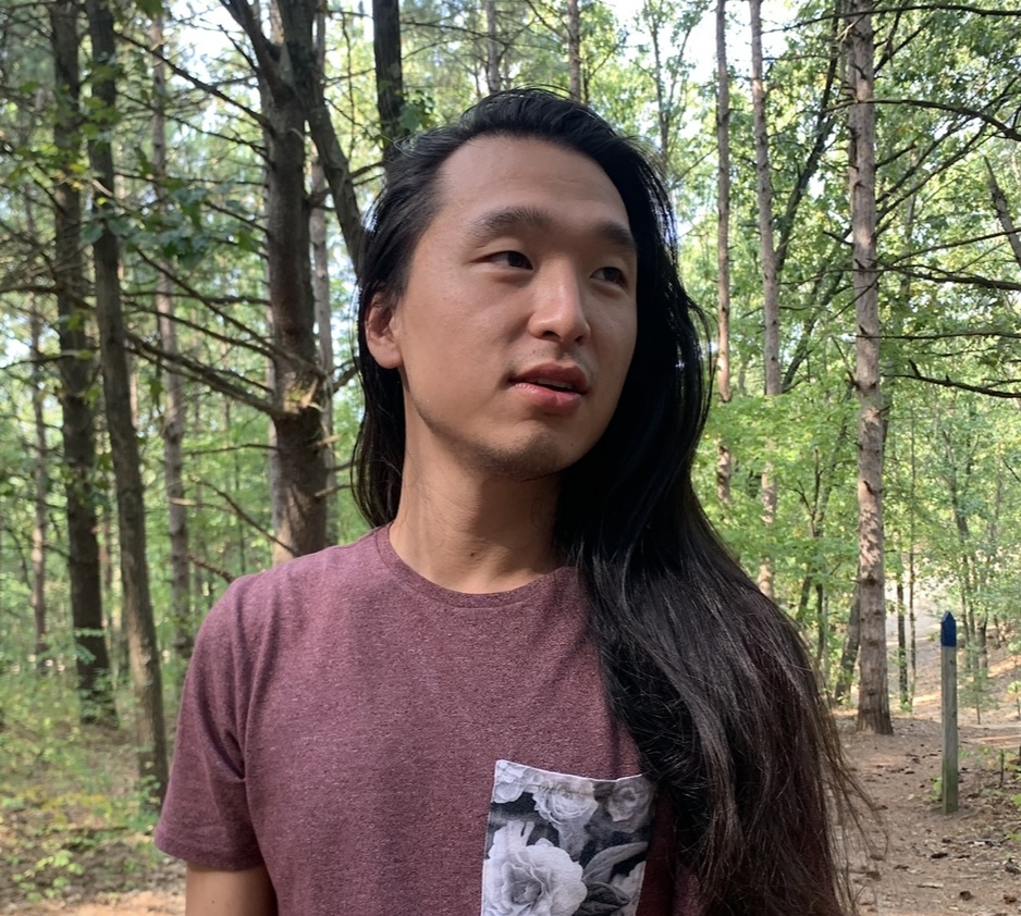 Man standing in the forest. He has long black hair and wears a mauve tshirt with a black-and-white pocket printed with roses. He is looking off to the side, his mouth open in a half-smile.