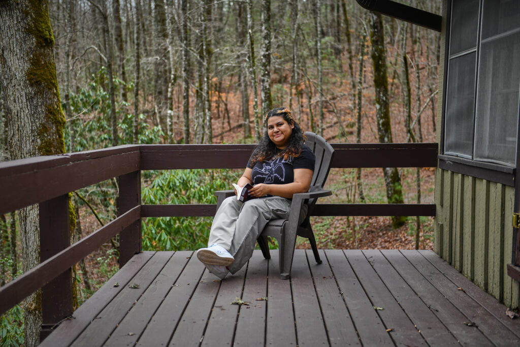 A curly-haired Indian woman sits on a grey chair. She is in a cabin deck in the mountains, with trees in the background. She is holding a book.