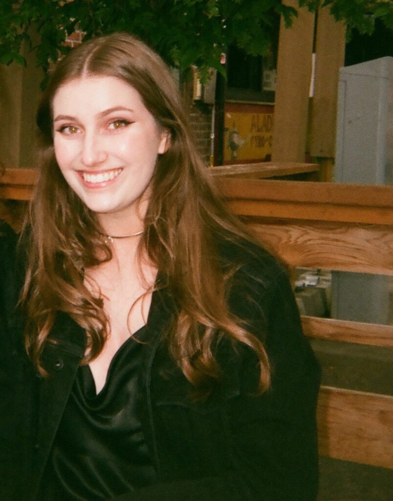 Sophie Johnson, a young woman with wavy brown hair, smiles at the camera. She is wearing a silver choker with a pendant and dark olive clothes. There is a tree and wooden structure behind her.