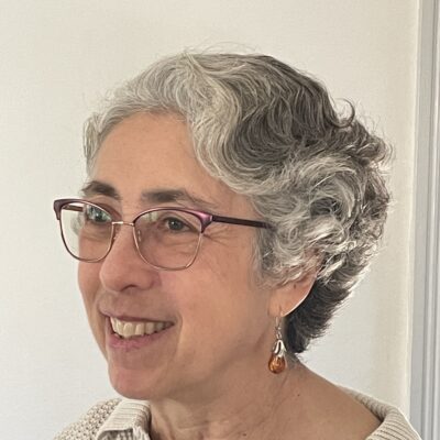Susanna Lang, a woman with short wavy white and gray hair, smiles, looking off to the side. She wears a knitted off-white collared shirt, glasses, and dangling earrings.
