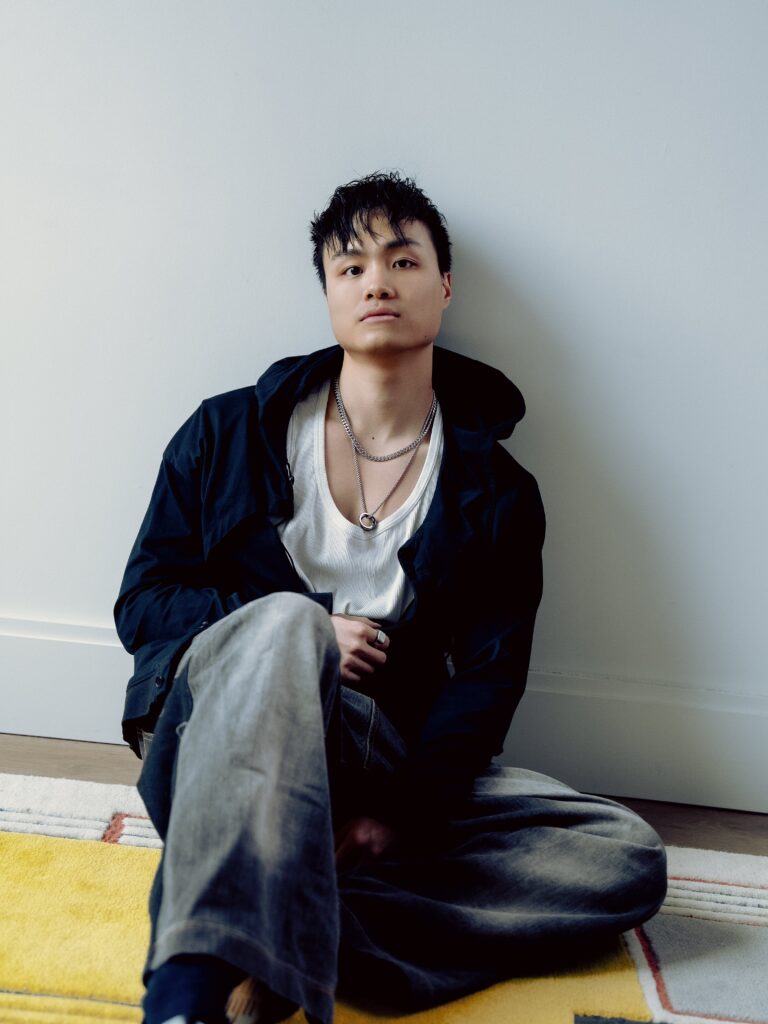 Tianyi, a poet with short black hair, sits on the floor on a yellow and white rug, leaning against a white wall, looking at the camera. He wears a loose white ribbed tank top underneath a midnight blue hoodie, faded jeans, and silver chains.