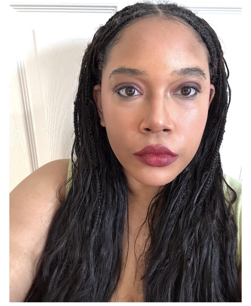 Tiezst "Tie" Taylor is a light-skinned Black femme wearing their hair in Boho Knotless braids with red lipstick and a green shirt against a white backdrop.
