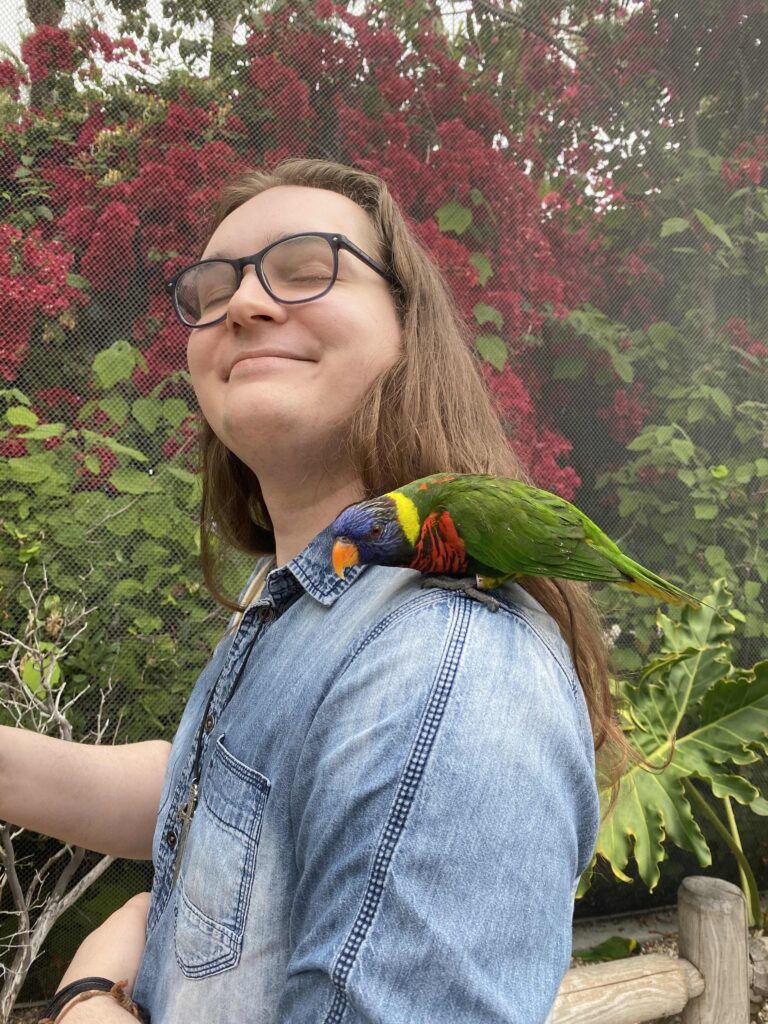 Pictured is James, a white nonbinary person with long brown hair and glasses. They are wearing a blue button-down denim shirt and a sword necklace, and have their eyes closed looking happily toward the sky. On their shoulder sits a colorful lorikeet; the background consists of red and green foliage. 
