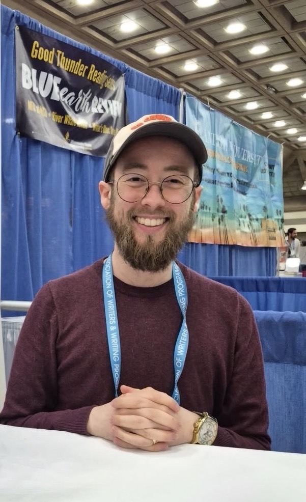 A white bearded man wearing a watch, a baseball cap, and a purple sweater sits at a booth with a blue backdrop.