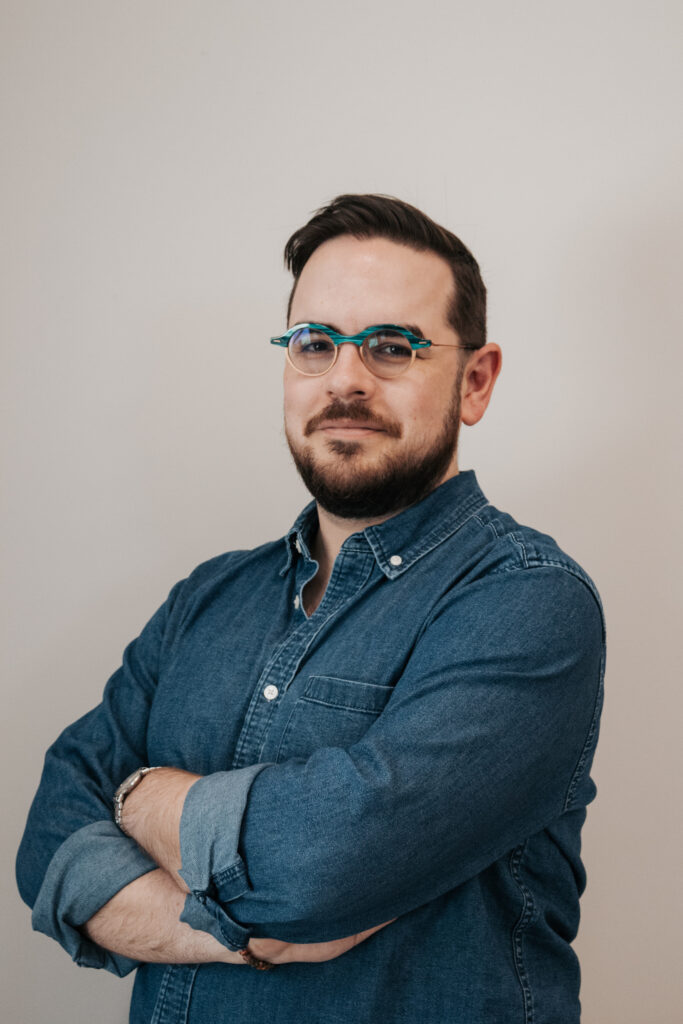 A half-length picture of Iain Grinbergs. He's smiling while standing with his arms crossed. He's wearing a denim shirt and round, mixed-material glasses.