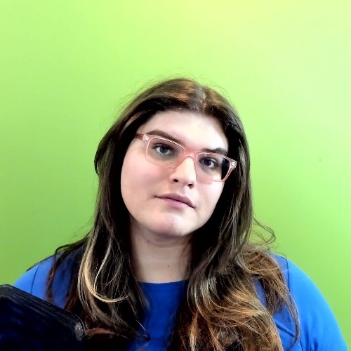 Sara Hovda, a transgender woman, stands against a bright green wall. She wears clear pink-rimmed glasses and a bright blue tshirt. She has long wavy brown hair.