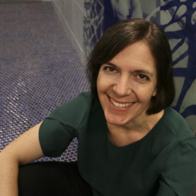 Smiling middle-aged white woman with brown eyes and dark brown hair in a bob with silver streaks, wearing a deep green, short-sleeved blouse and sitting on a blue-tiled floor, with oceanic blue and gray wall tile behind her.