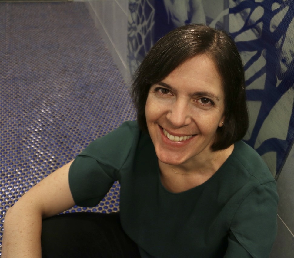 Smiling middle-aged white woman with brown eyes and dark brown hair in a bob with silver streaks, wearing a deep green, short-sleeved blouse and sitting on a blue-tiled floor, with oceanic blue and gray wall tile behind her.