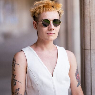 A person with short, ruffled blond hair stands near a stone column, wearing round sunglasses and a white tailored vest. They have several American traditional tattoos on both arms and a neutral expression.