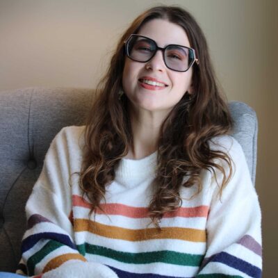 Elina Katrin, a poet with long wavy brown hair, smiles widely on a grey sofa against a soft beige wall. She wears a white sweater with horizontal stripes and large tinted black-framed glasses.