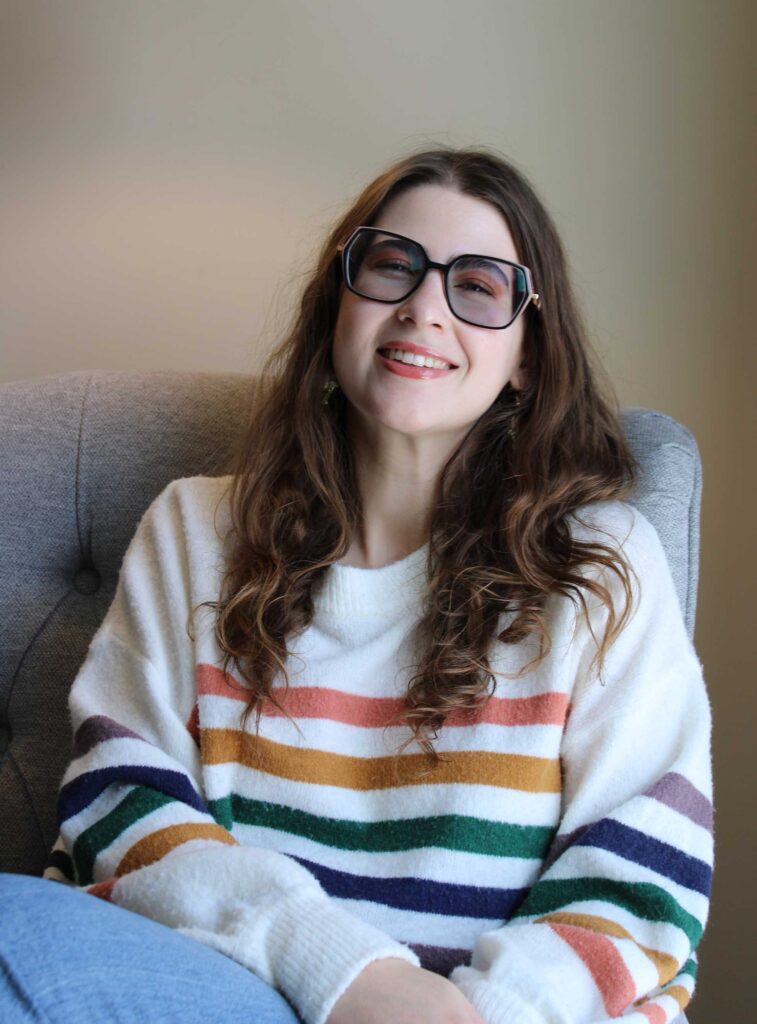 Elina Katrin, a poet with long wavy brown hair, smiles widely on a grey sofa against a soft beige wall. She wears a white sweater with horizontal stripes and large tinted black-framed glasses.