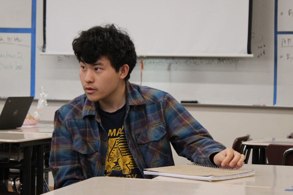 Ethan Kwak, a young Asian American poet with thick wavy black hair, sits in a classroom, looking off to the side. He wears a black tshirt with deep yellow graphic underneath a blue-and-brown plaid shirt. There is a book on the desk to his left and white sheets of paper and a whiteboard on the wall behind him. 