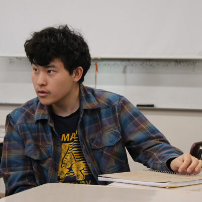 Ethan Kwak, a young Asian American poet with thick wavy black hair, sits in a classroom, looking off to the side. He wears a black tshirt with deep yellow graphic underneath a blue-and-brown plaid shirt. There is a book on the desk to his left and white sheets of paper and a whiteboard on the wall behind him.