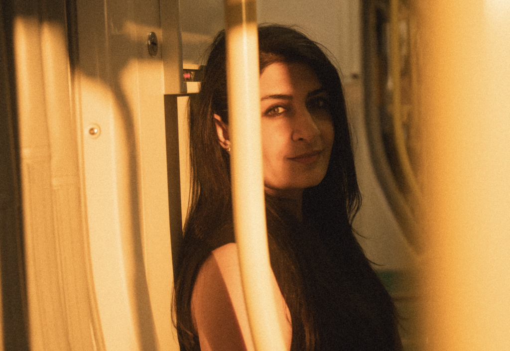 Hananah Zaheer, a South Asian woman, stands on a train, looking at the camera. There is a thin pole between her and the camera and golden sunlight cast on her. She has long black hair and is wearing a sleeveless black top.