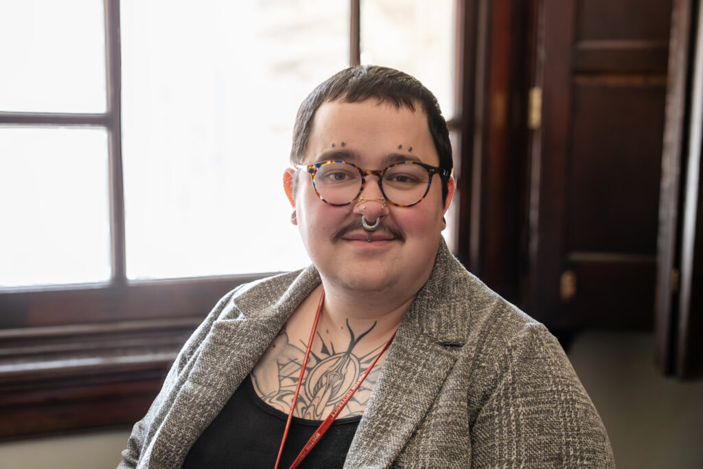A photo of Sterling-Elizabeth, a white trans woman with short black hair, sitting in front of a bright window, showing only her upper torso and head. She is heavily pierced, her chest is prominently tatooed, she has glasses and a mustache, and she is wearing a gray jacket over a black dress.